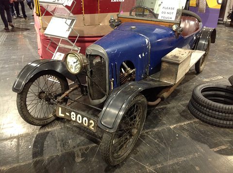 A weathered blue cyclecar with a mesh radiator, single headlamp and black wings, a stack of tyres beside it. Opens a larger view