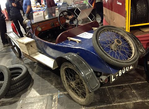 Rear quarter view of a weathered blue cyclecar with a wooden box and a spare wire wheel on its tail. Opens a larger view