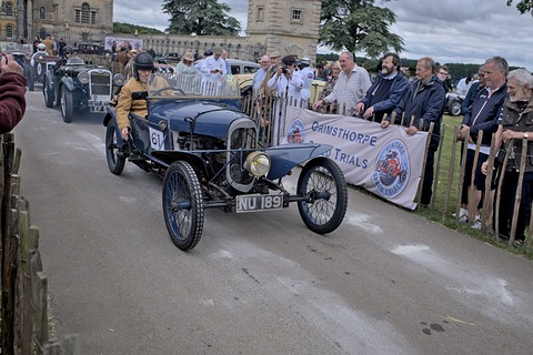 A helmeted driver in a blue cyclecar on a drive, spectators behind a fence and a Grimsthorpe Trials banner. Opens a larger view