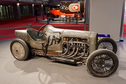 Side view of an unpainted racing car with a long bonnet, exposed engine and four exhaust pipes. Opens a larger view