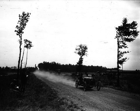A small dark racing car trailing a long plume of dust along a straight dirt road lined with tall thin trees. Opens a larger view