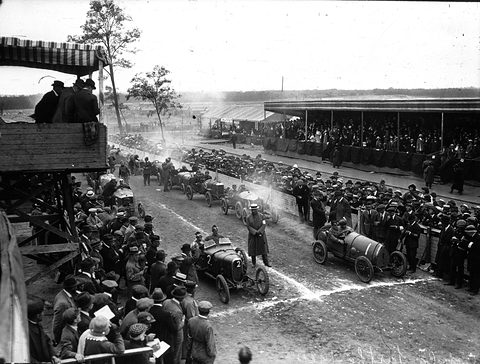 Seen from a timber stand, small racing cars lined up on a dirt road between packed crowds, a covered grandstand beyond. Opens a larger view