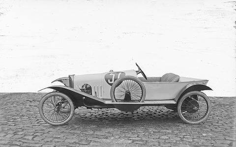Side view of a pale two-seat cyclecar with a spare wheel on its flank and a pointed tail, on cobbles against a blank sky.