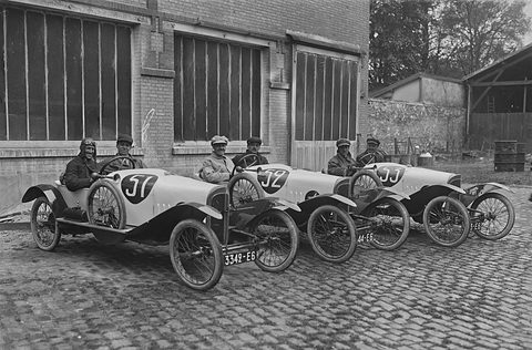 Three pale cyclecars with large black numbers on their bonnets lined up on cobbles, two men in each, brick hangar behind. Opens a larger view