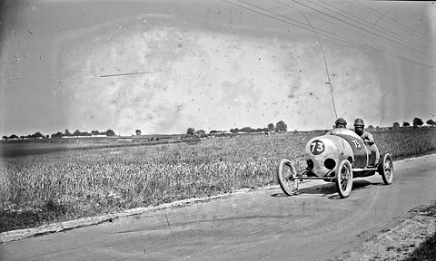 A small pale racing cyclecar numbered 73 rounds a bend on a country road beside a cornfield. Opens a larger view