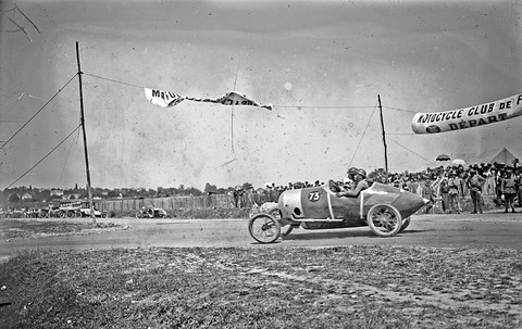 A pointed-tail racing cyclecar on a road past a crowd behind a fence, banners strung overhead. Opens a larger view