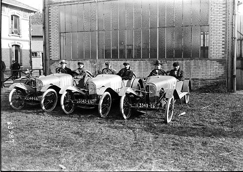 Three small open cyclecars with pointed noses parked side by side on grass, each with driver and passenger in caps. Opens a larger view