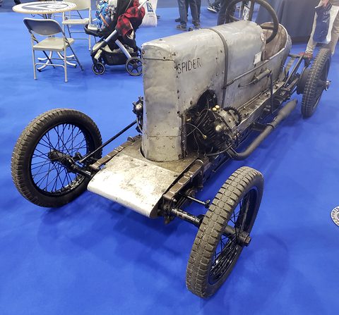 A narrow unpainted sprint car with a tall bonnet lettered Spider, exposed engine and wire wheels on blue carpet.