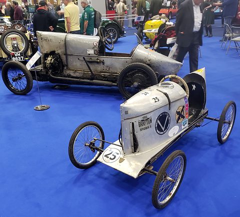 A bare-metal sprint special behind a small silver soapbox car numbered 25, on a blue carpet. Opens a larger view