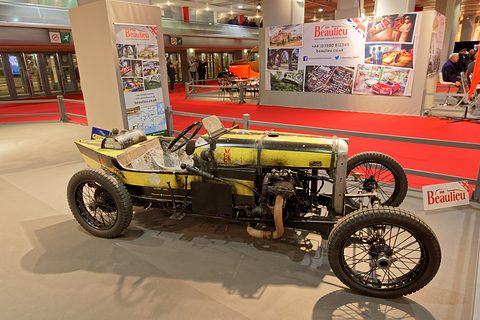 A yellow and black racing special with an exposed engine and wire wheels on a grey stand before Beaulieu banners. Opens a larger view
