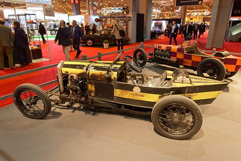 Side view of a yellow and black racing special with an exposed engine, visitors and other cars behind. Opens a larger view