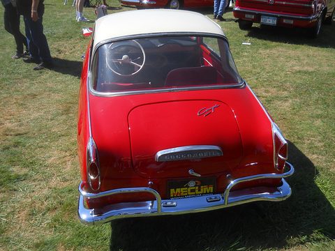Rear view of a red coupé with a white roof on grass with an auction plate on the bumper. Opens a larger view