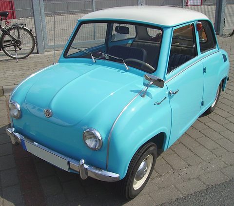 Bright light-blue saloon with a white roof parked on paving beside a railing, seen from the front left. Opens a larger view