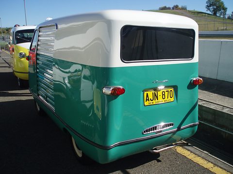 Rear three-quarter of a teal and white van with large rear window, on a car park. Opens a larger view