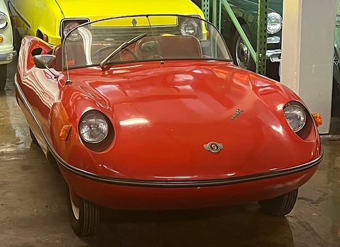 Orange open roadster in a museum, round headlamps in curved wings, a yellow car behind. Opens a larger view