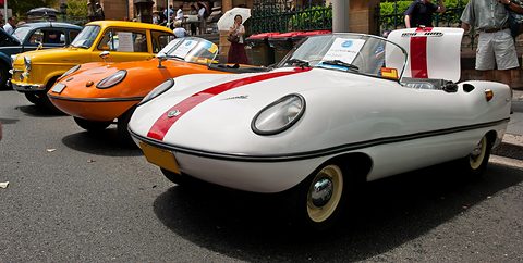 White open roadster with a red centre stripe, an orange and a yellow car beside it and visitors behind. Opens a larger view