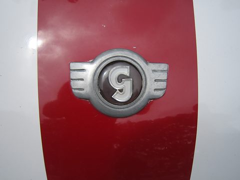 A chrome winged badge with a G on a red stripe running over white paint. Opens a larger view