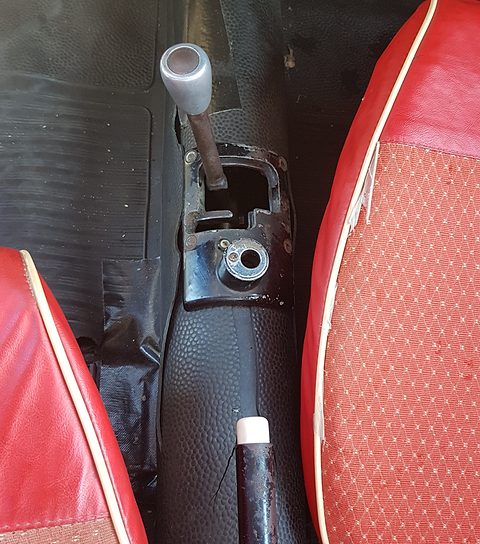 Chrome gear lever rising from a rubber boot between two red patterned seats. Opens a larger view