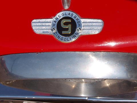 Chrome winged badge with a G in the centre on red paint above a chrome bumper. Opens a larger view