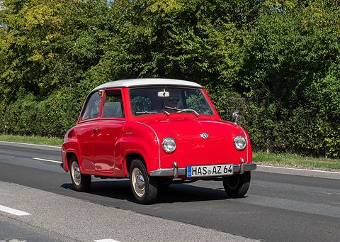 Red saloon with a white roof driving along a country road in front of a hedge. Opens a larger view