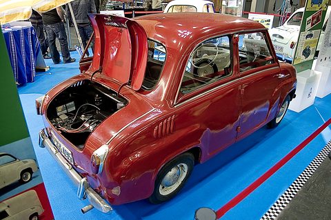 Red saloon seen from the rear left with its engine cover raised, on a blue show floor. Opens a larger view