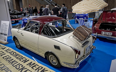 Cream coupé with a dark red roof and a wicker hamper on its rack, on a blue show carpet. Opens a larger view