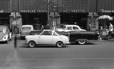 White coupé at the kerb in front of a newspaper building, an American saloon and a Volkswagen nearby