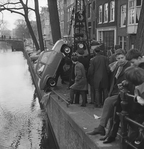 Small saloon tipped nose-up against a crane on a canal bank, onlookers crowding the railings beside the water