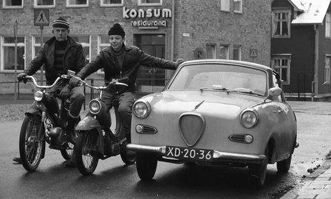 Two young men on mopeds pose beside the nose of a small coupé on a wet street in front of a restaurant sign. Opens a larger view