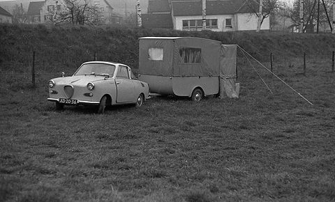 Small coupé parked beside a trailer with a canvas tent folded out from it, on grass by a fence. Opens a larger view