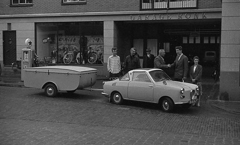 Pale coupé and flat trailer at a kerb outside a garage with a petrol pump, several men in suits watching. Opens a larger view