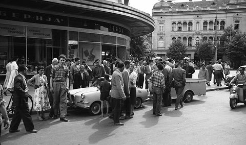 Crowd of men and boys gathered round a small pale coupé and trailer on a city square in front of shops. Opens a larger view