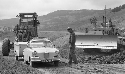 Pale coupé towing a box trailer on a muddy track beside a road grader and a bulldozer, a workman standing by. Opens a larger view