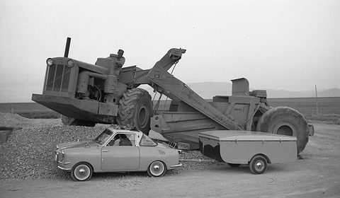 Tiny coupé and trailer parked in front of a huge wheeled earth-moving scraper on open ground. Opens a larger view