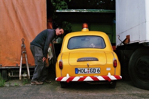 Yellow saloon seen from behind with a red and white striped bumper, a man reaching into a lorry beside it. Opens a larger view