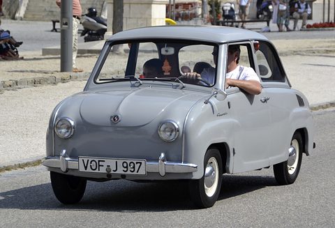 Grey saloon driving towards the camera with two occupants, spectators along the road. Opens a larger view