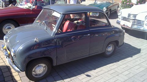 Dark blue saloon parked on paving in sunshine, seen from the side with a red interior visible. Opens a larger view