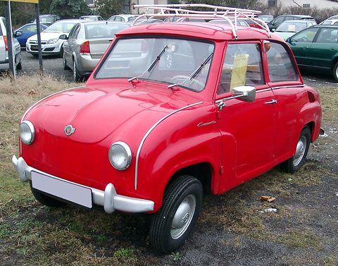 Red saloon with a chrome roof rack parked on rough grass, seen from the front right. Opens a larger view