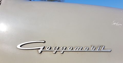 Close-up of a chrome script badge reading Goggomobil on pale grey paint, a strip of blue sky above. Opens a larger view