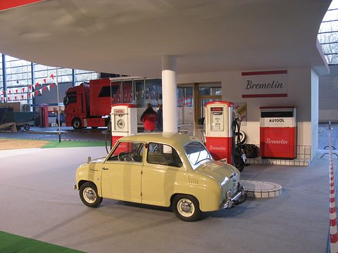 Yellow saloon beside red and white petrol pumps under a canopy, in a reconstructed period scene. Opens a larger view