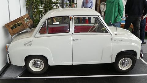 White two-door saloon with red seats, side view, a wooden crate on a stand behind. Opens a larger view