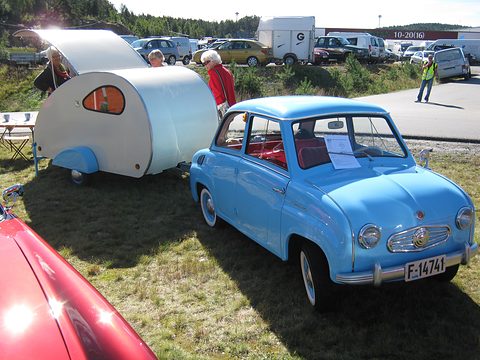Sky-blue saloon beside a small silver teardrop caravan on grass at an outdoor show. Opens a larger view