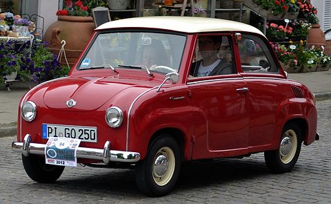 Red saloon with a white roof being driven on cobbles, seen from the front left, the driver visible. Opens a larger view
