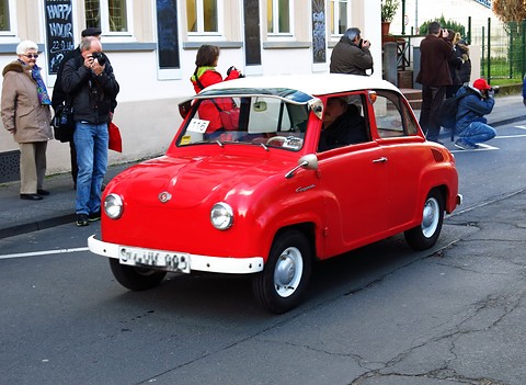 Red saloon with a white roof on a street, spectators and photographers behind it. Opens a larger view