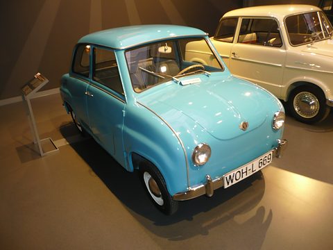 Light blue saloon under spotlights in a dark museum hall, a cream car behind. Opens a larger view