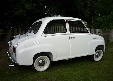 White saloon with whitewall tyres on grass, seen from the rear left in front of a wooden fence. Opens a larger view