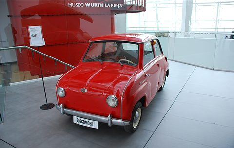 Red saloon with a white Goggomobil sign at its bumper on a grey floor in a modern museum hall