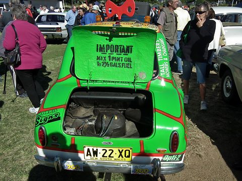 Green car covered in sponsor lettering seen from behind with its engine cover up, people around it on grass. Opens a larger view
