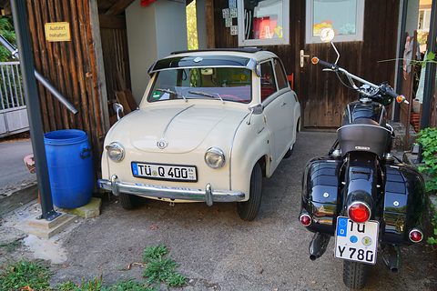 Cream saloon seen from the front parked in a yard beside a black motorcycle and a blue barrel. Opens a larger view