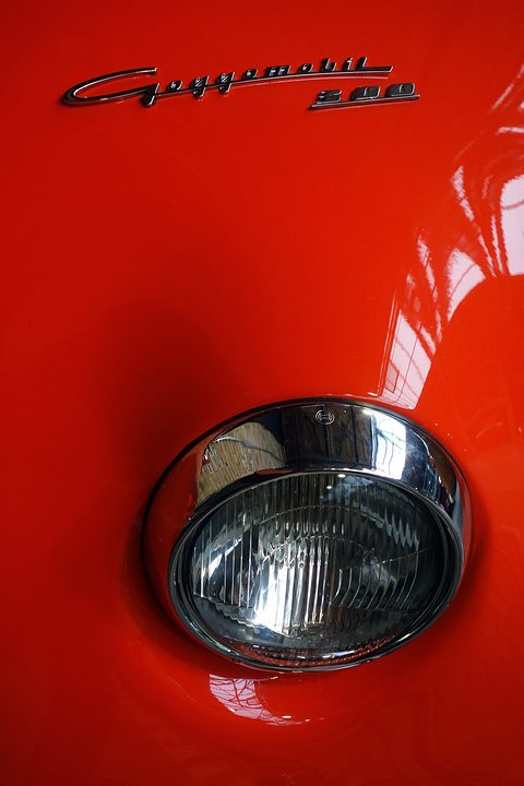 Close-up of a chrome headlamp below a Goggomobil script badge on red paint. Opens a larger view